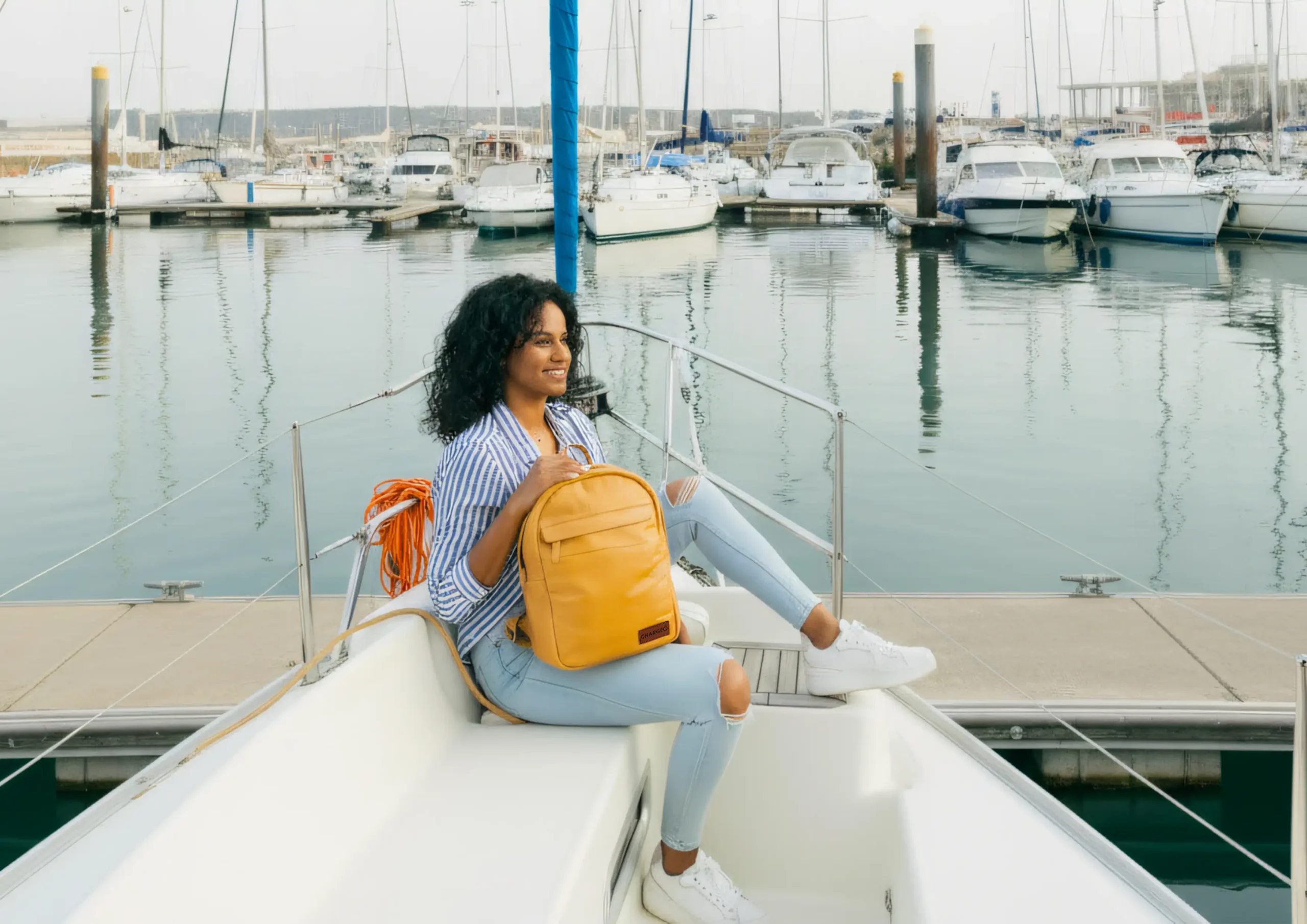Woman wearing ripped jeans and a striped shirt sitting on a yacht, holding the CHARGEO Butter Tan leather
