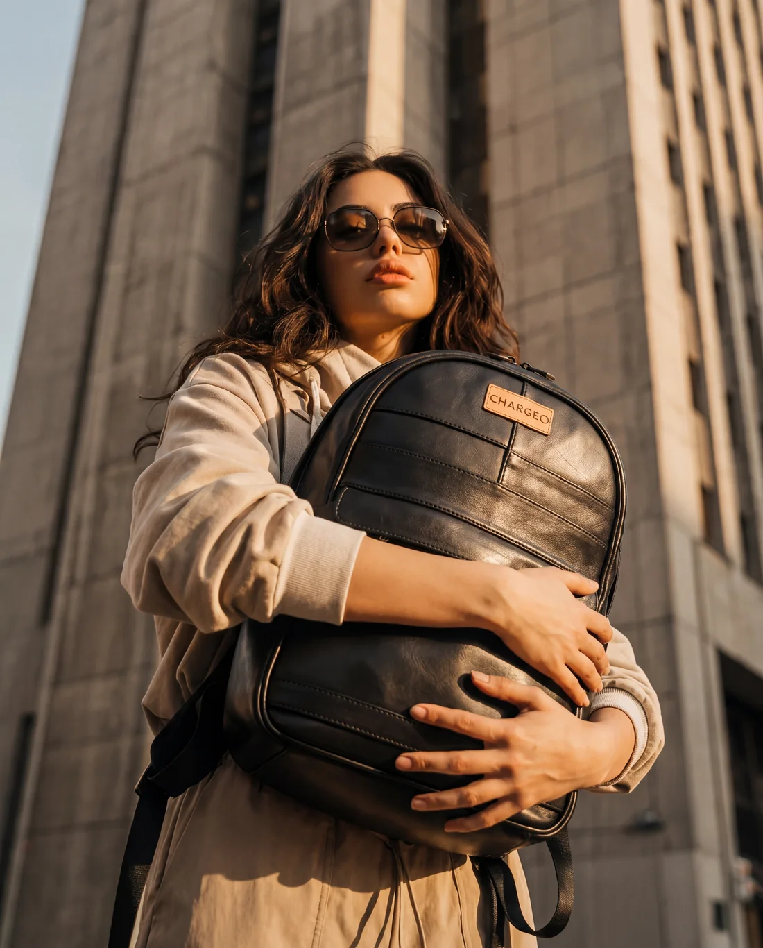 Woman in sunglasses holding a CHARGEO charcoal leather backpack in front of a city tower