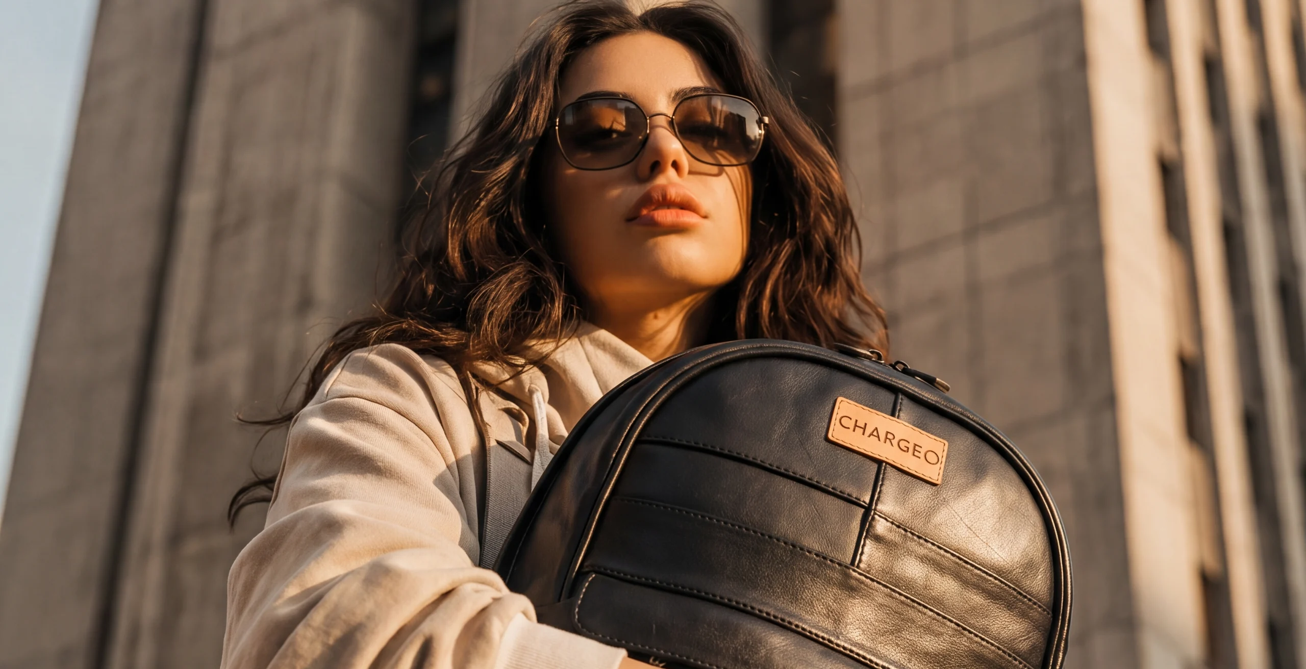 Woman in sunglasses holding a CHARGEO charcoal leather backpack in front of a city tower