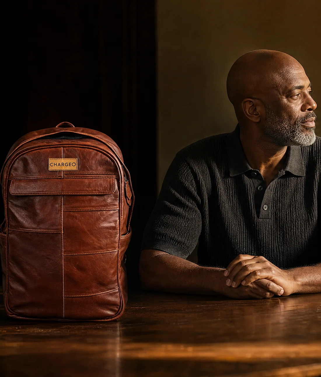 Man seated at an oak table in a library with a CHARGEO cognac leather backpack standing on the table