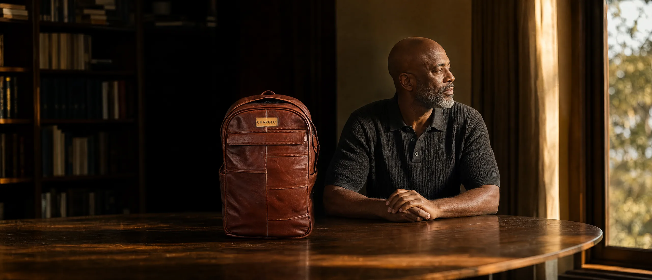 Man seated at an oak table in a library with a CHARGEO cognac leather backpack standing on the table