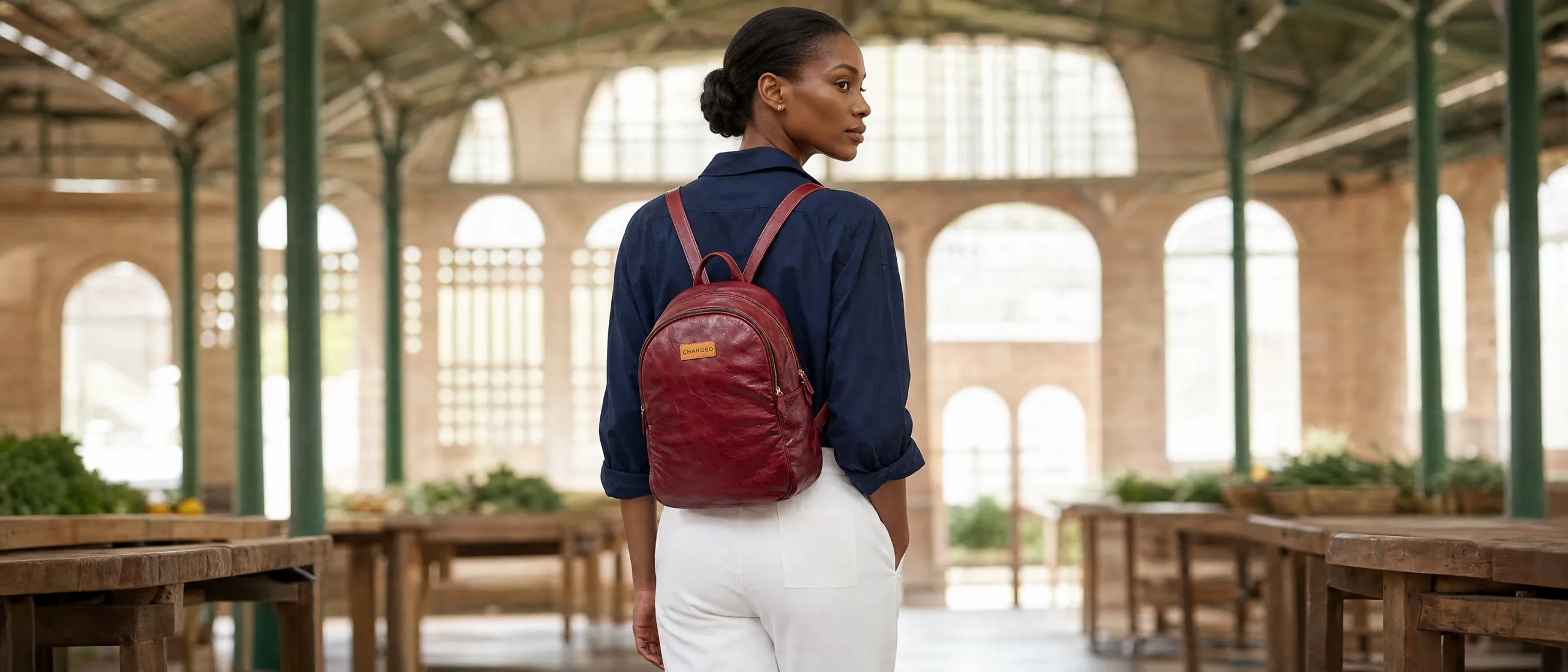 Lady in a navy shirt and white trousers wearing a red CHARGEO leather backpack on both shoulders in a market hall