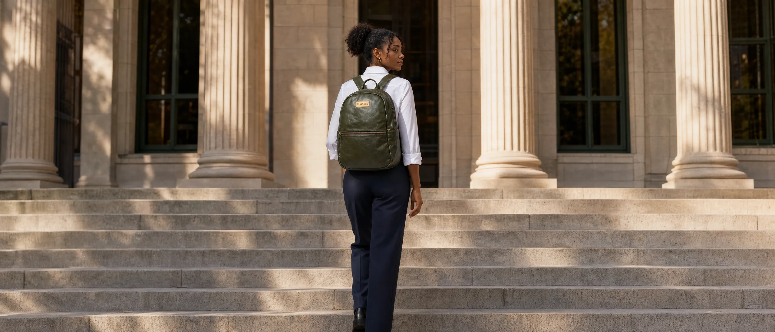 Lady in a white shirt and navy trousers wearing a green CHARGEO leather backpack on both shoulders, walking up stone steps