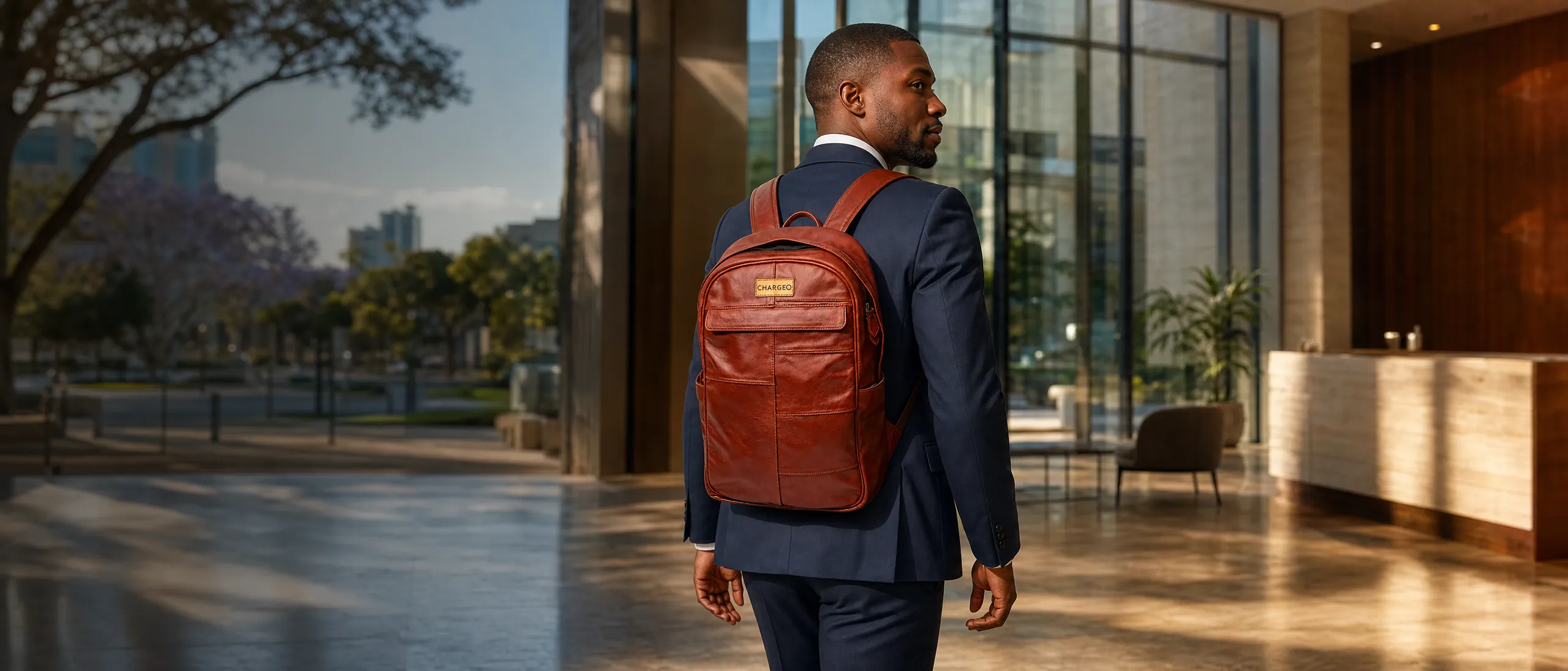 Man in a navy suit walking into an office reception in Rosebank wearing the CHARGEO Stellenbosch Leather Backpack in Cognac on both shoulders