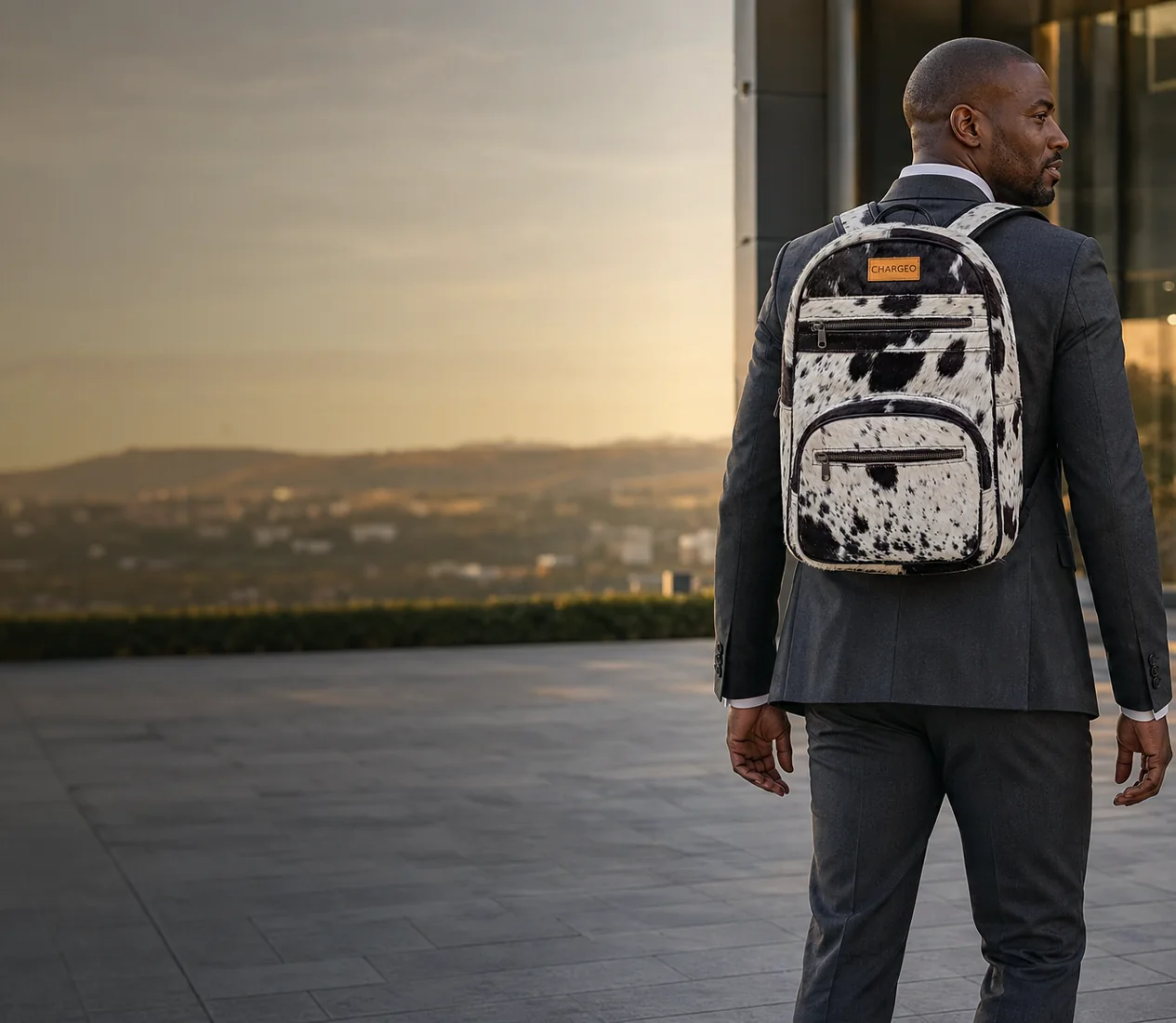 Man in a charcoal suit leaving an office tower in Sandton at golden hour wearing the CHARGEO Zulu Treasure Leather Backpack in black and white Nguni hide