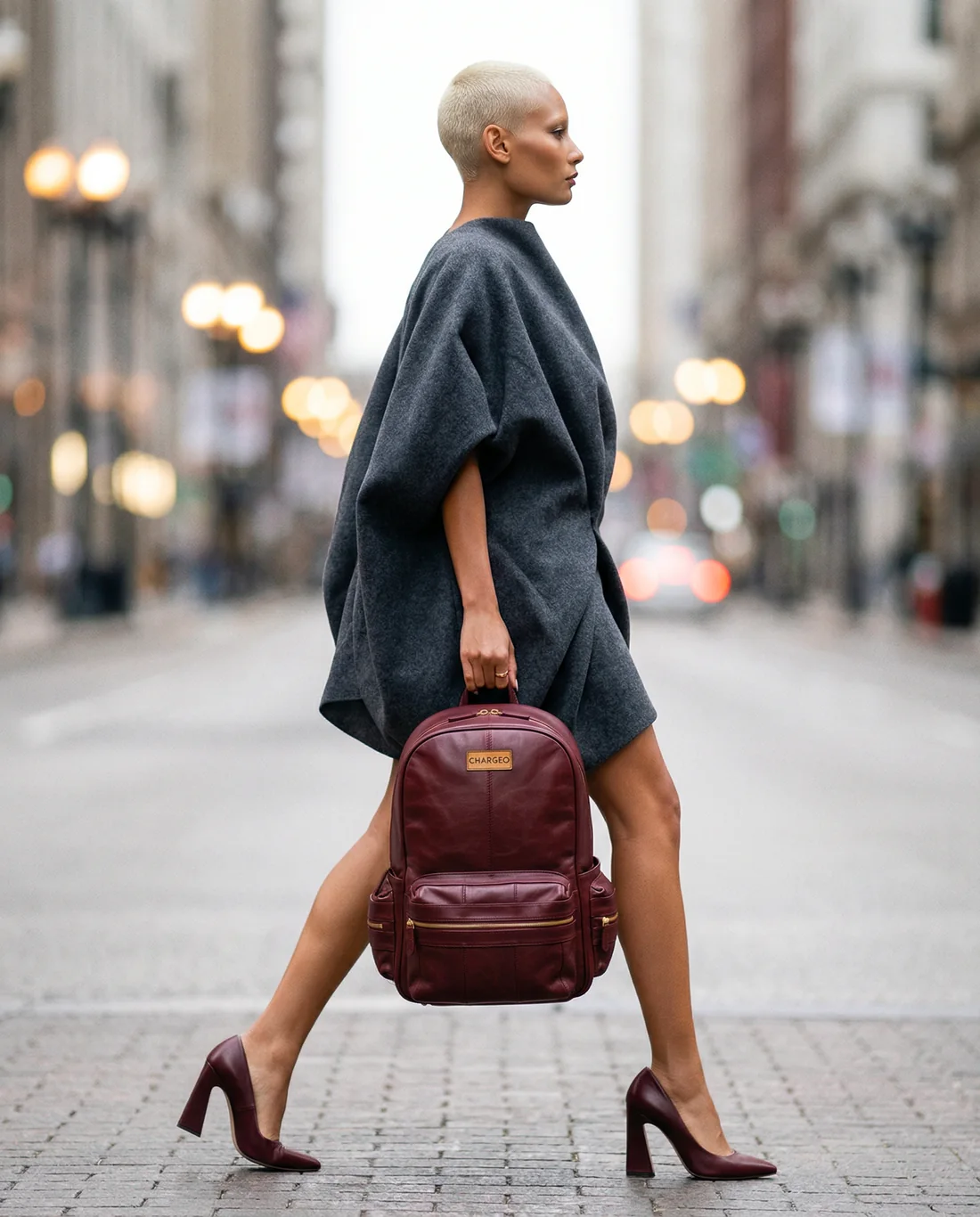 Woman walking through a city street carrying a CHARGEO burgundy leather backpack by its handle