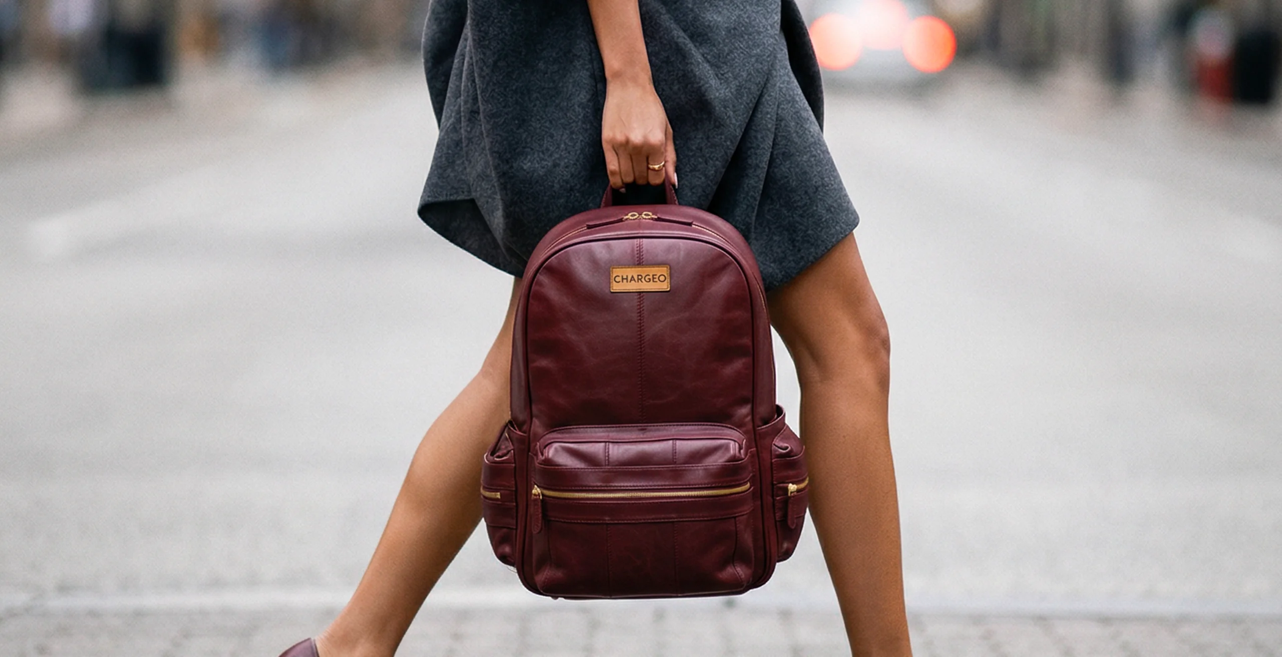 Woman walking through a city street carrying a CHARGEO burgundy leather backpack by its handle