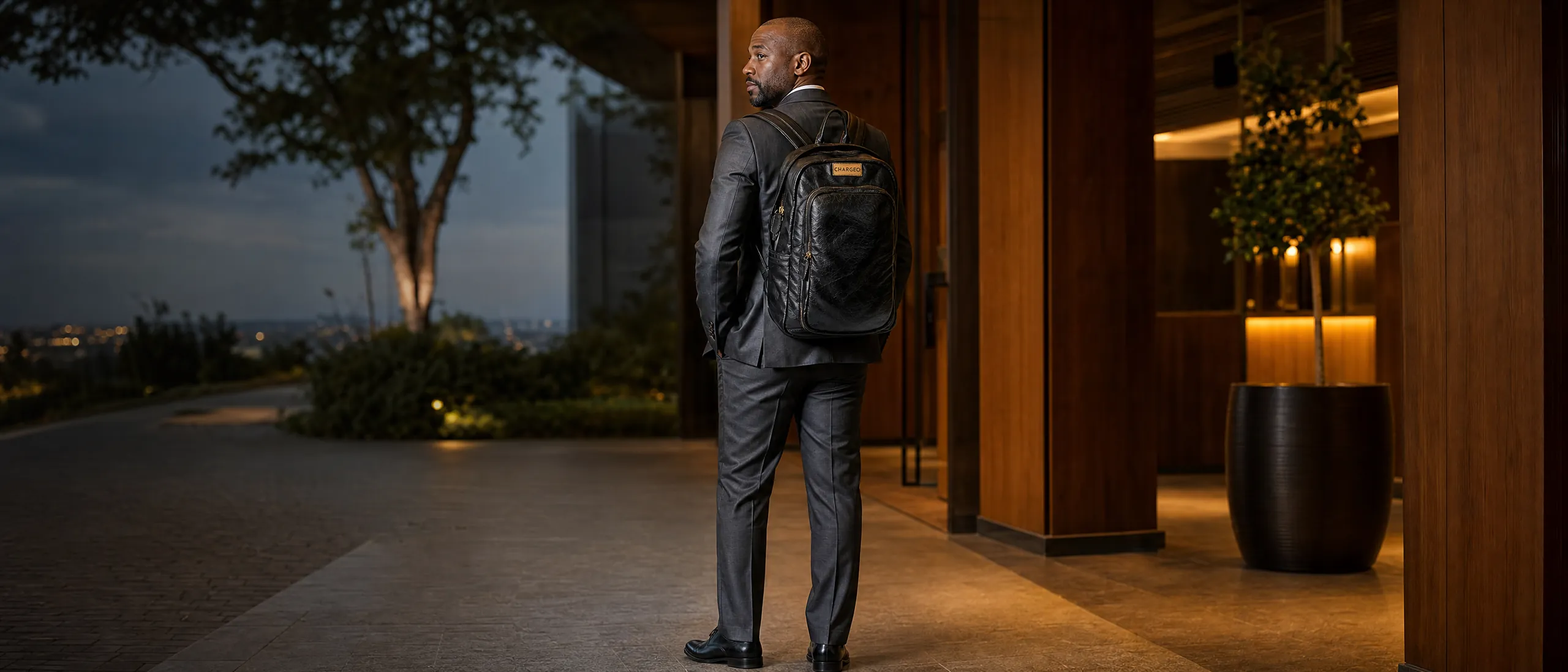 Man in a charcoal three-piece suit in the wood-panelled lobby of a Sandton hotel wearing the CHARGEO Parktown Leather Backpack in Charcoal on both shoulders