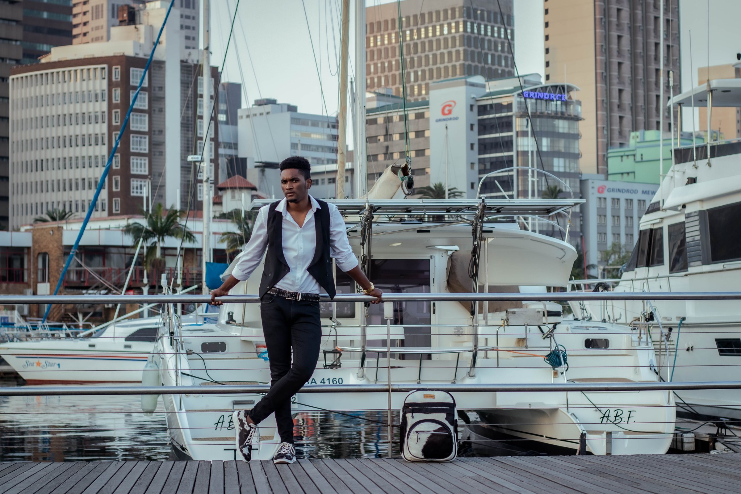A man standing on the marina boardwalk in Durban with the CHARGEO black and white hide backpack at his feet, boats and city towers behind him