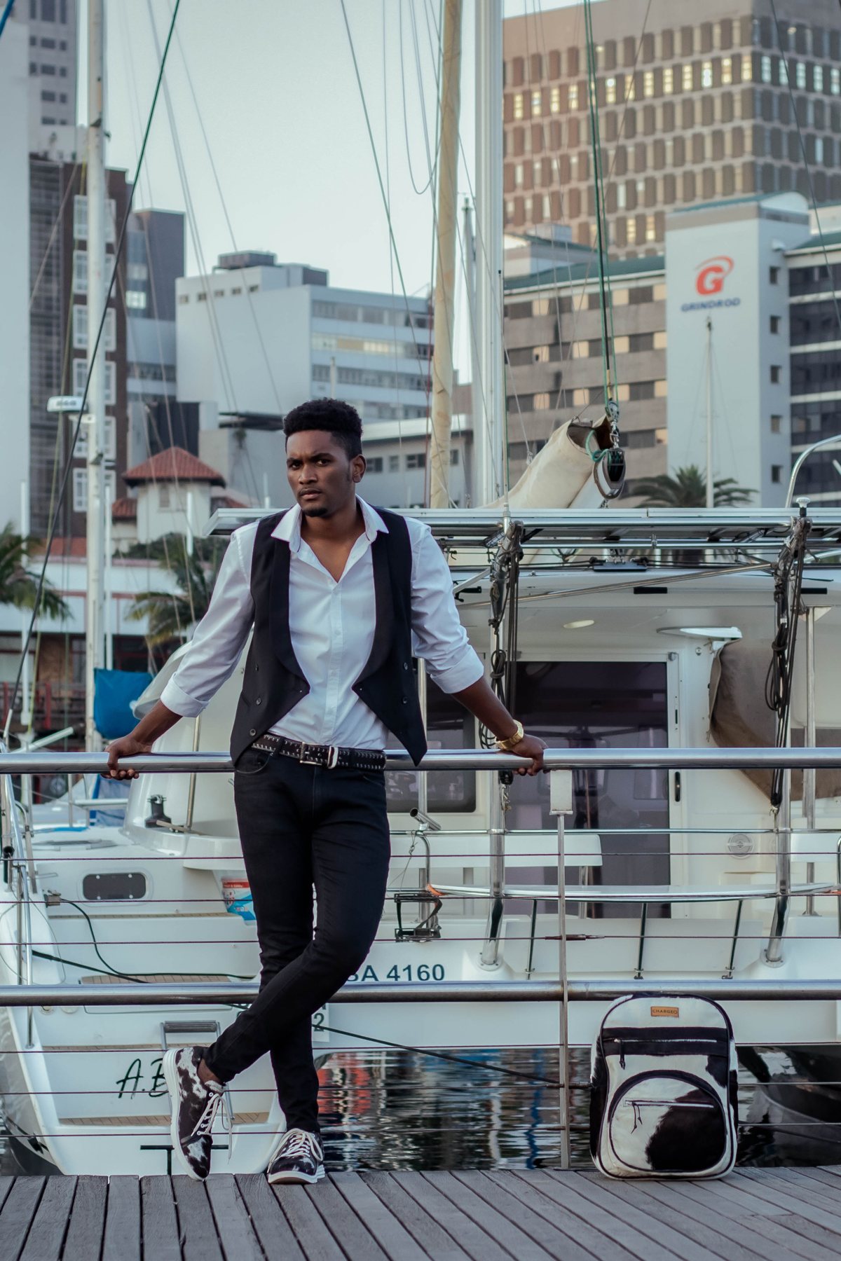A man standing on the marina boardwalk in Durban with the CHARGEO black and white hide backpack at his feet, boats and city towers behind him