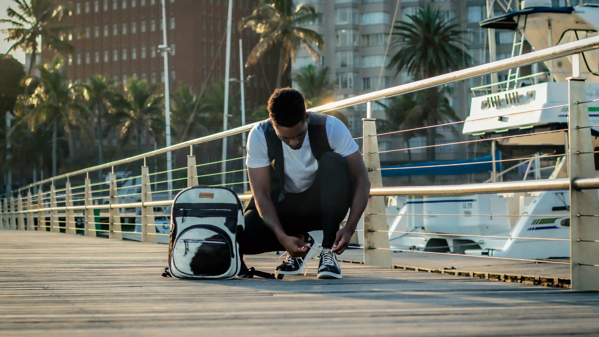 A man crouching on the marina boardwalk to tie his shoe beside the CHARGEO black and white hide backpack, August 2023