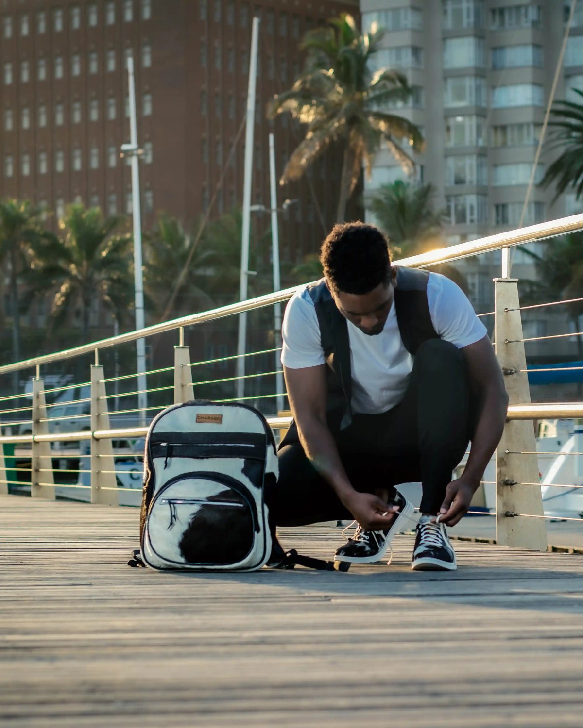 A man crouching on the marina boardwalk to tie his shoe beside the CHARGEO black and white hide backpack, August 2023