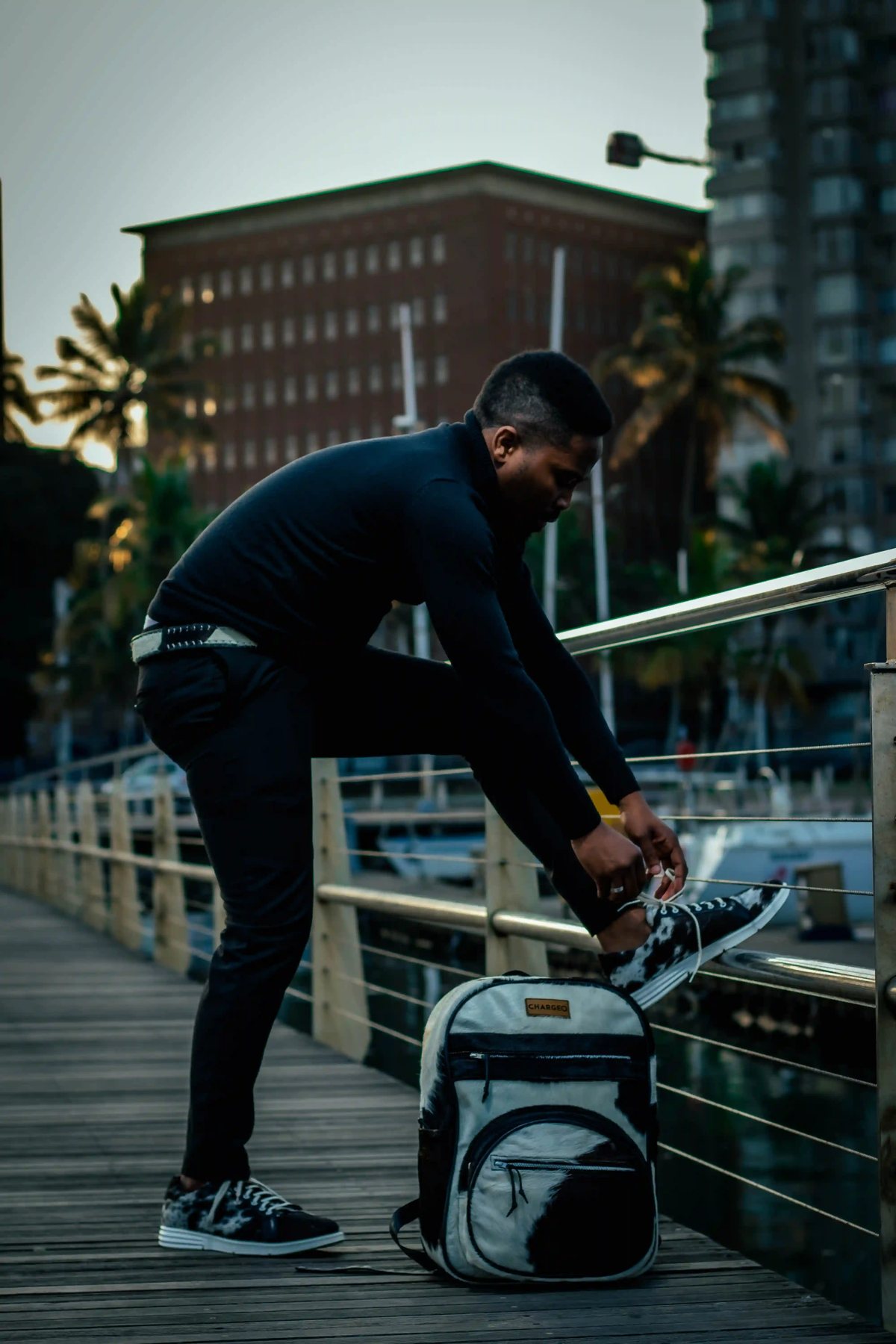 A man in black tying his shoe at the marina rail beside the CHARGEO black and white hide backpack, August 2023